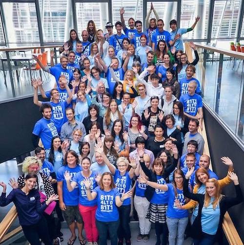 A large group of mostly women who attended the Django Girls free programming workshop in Ljubljana. The workshop attendees and mentors are posing on stairs and smiling and waving at the camera.